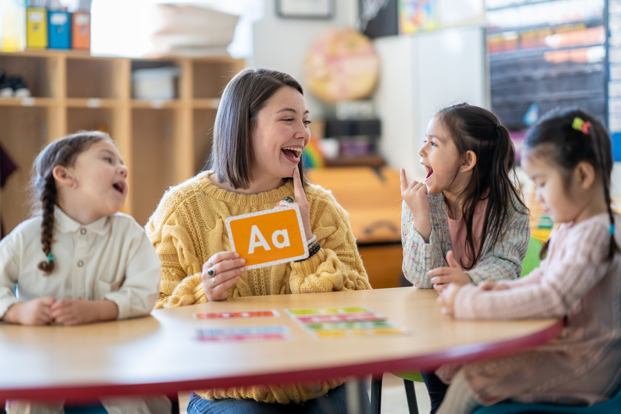 A young woman appearing to be a teacher with light skin and short dark hair is sitting at a table with 3 young school-aged girls. The woman is holding up a flashcard with the letter A on it, while the girls look at it.