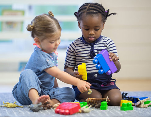 Two young girls, one with light skin and blonde hair in pigtails, the other with dark skin and brown hair with braids, play with large blocks on the carpet.