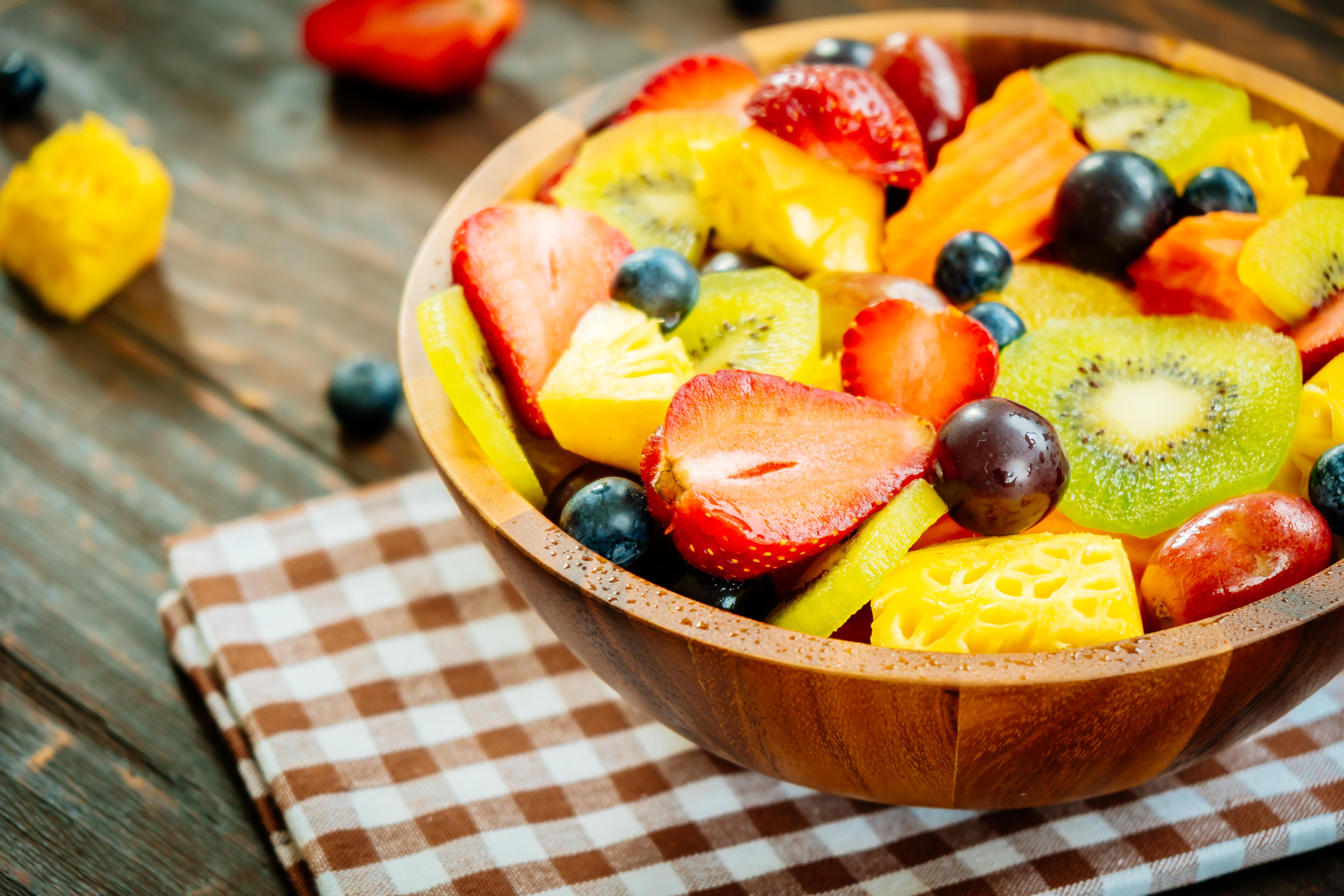 A bowl of Mixed Assorted Fruits including strawberries, blueberries, pineapple, and kiwi. The bowl is sitting on a wooden table with a checkered pattern table cloth underneath it. 