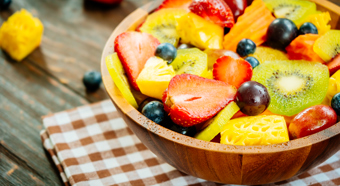 A bowl of Mixed Assorted Fruits including strawberries, blueberries, pineapple, and kiwi. The bowl is sitting on a wooden table with a checkered pattern table cloth underneath it. 