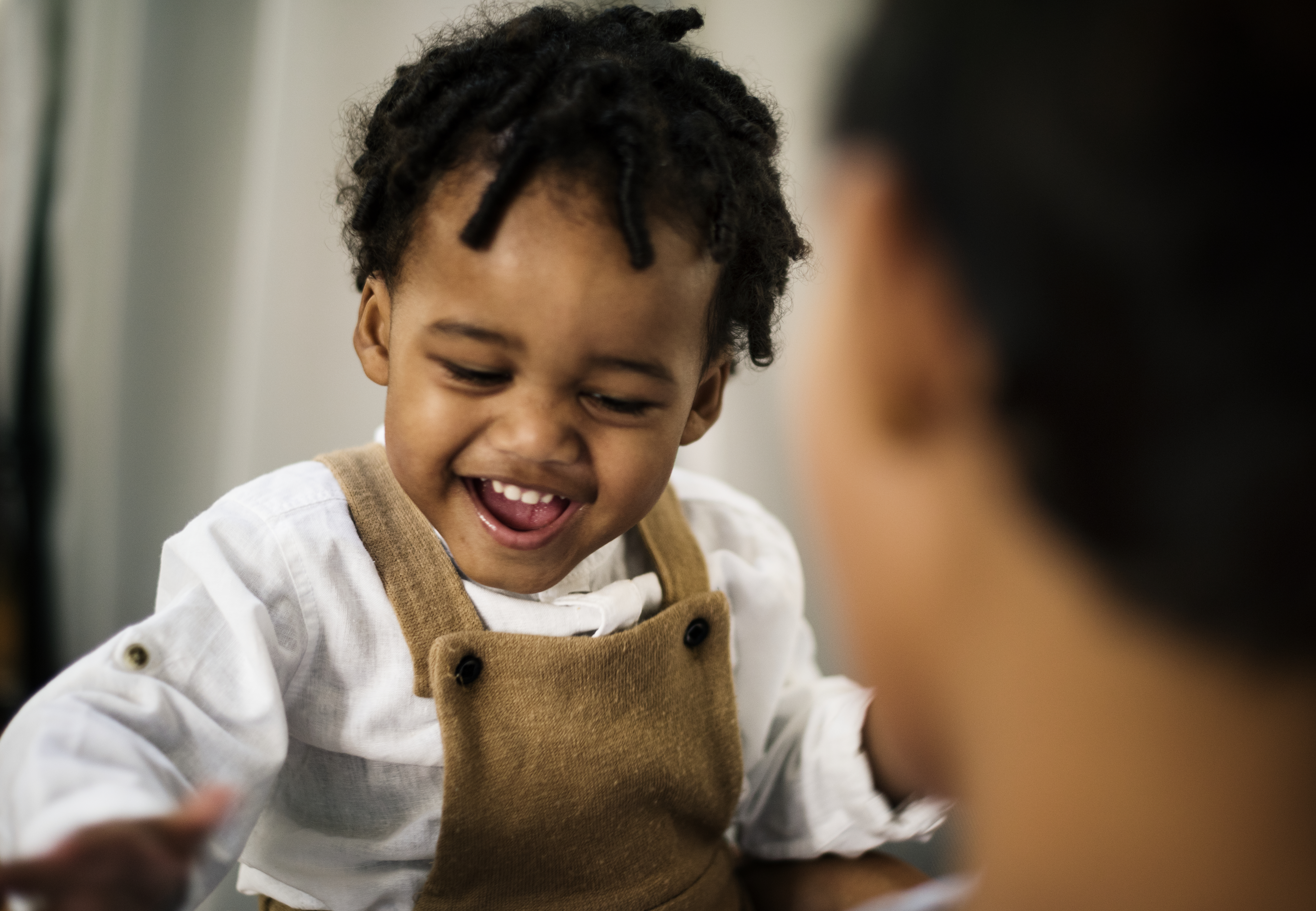 Closeup of African American toddler boy laughing and smiling, wearing brown overalls with a white shirt.