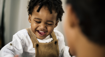 Closeup of African American toddler boy laughing and smiling, wearing brown overalls with a white shirt.