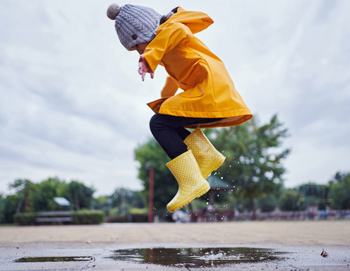 A young girl dressed in a yellow raincoat and boots is jumping in a puddle.