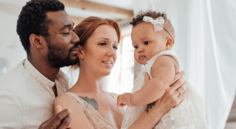 Adult male and female smile and look at baby that the female is holding in front of her. Baby is dressed in a white dress. Indoor bright setting.