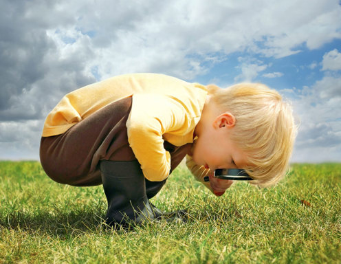 A toddler-aged boy with light skin and blonde hair is bending over looking at the grass with a magnifying glass.