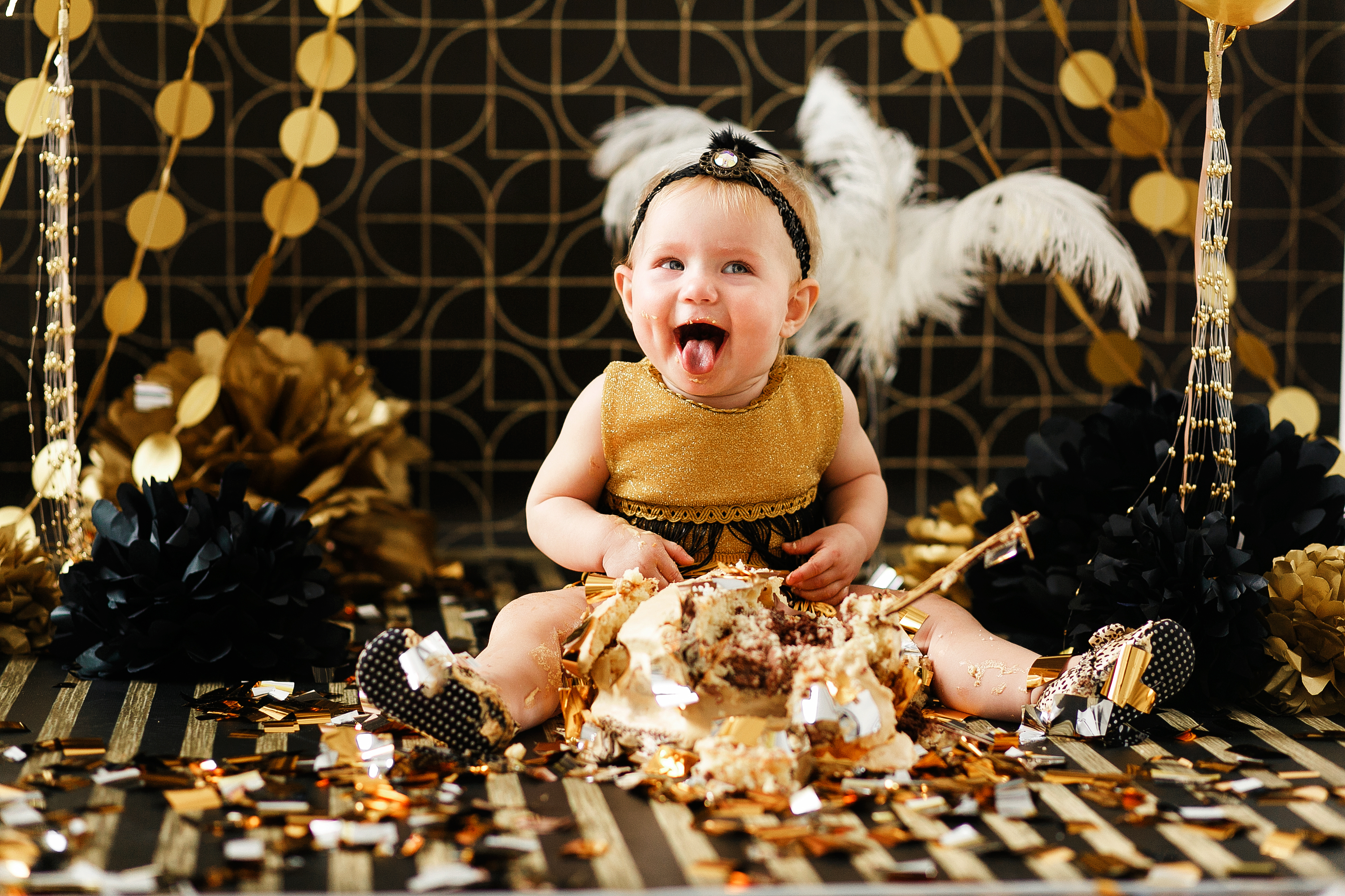 Happy baby wearing sparkly gold and black clothing smiles with their tongue out while eating some kind of cake. New years eve decorations in background. 