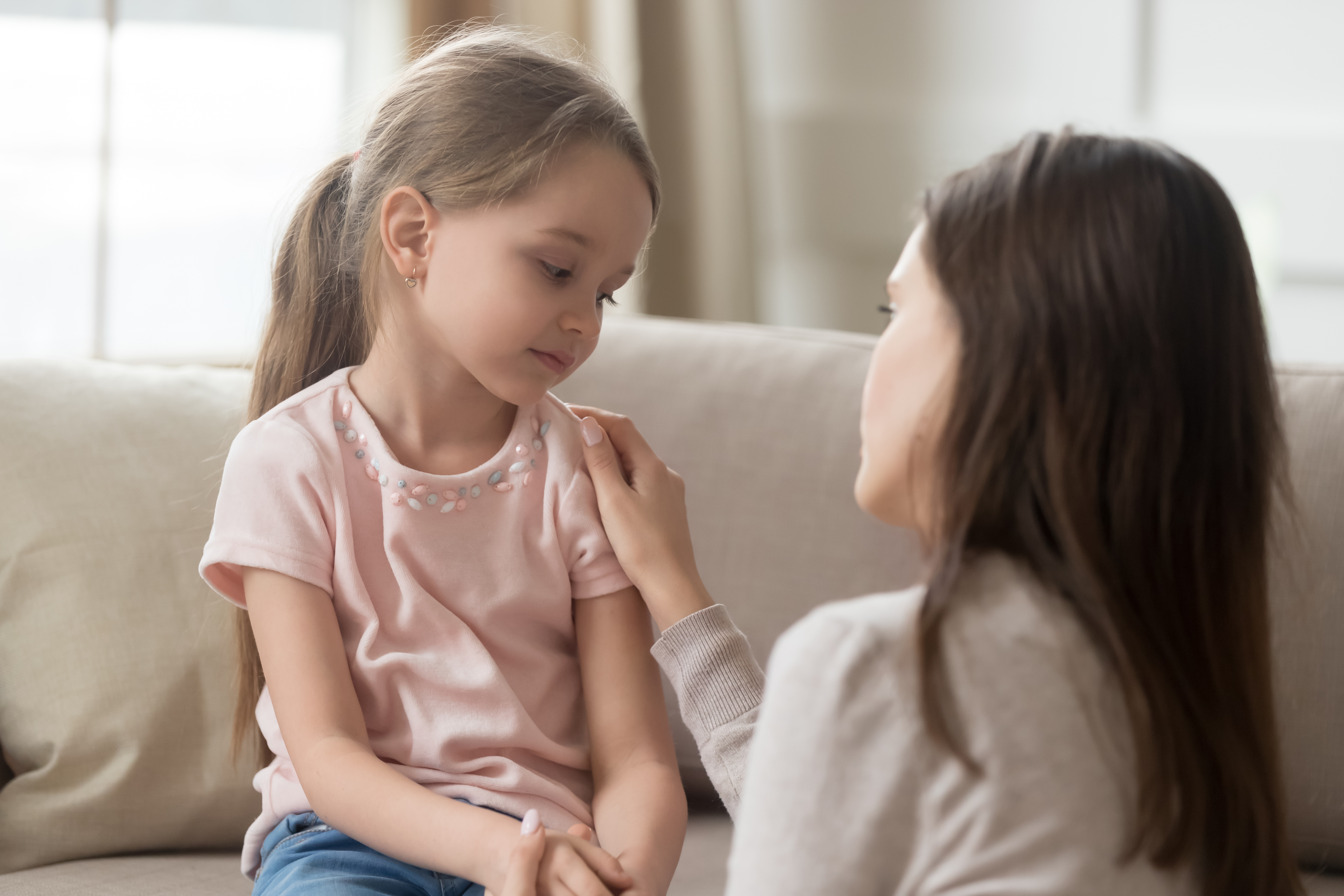 Girl wearing pink shirt sitting on couch with a sad looking expression, while adult female with her hand on the child's shoulder speaks to her.