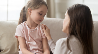 Girl wearing pink shirt sitting on couch with a sad looking expression, while adult female with her hand on the child's shoulder speaks to her.