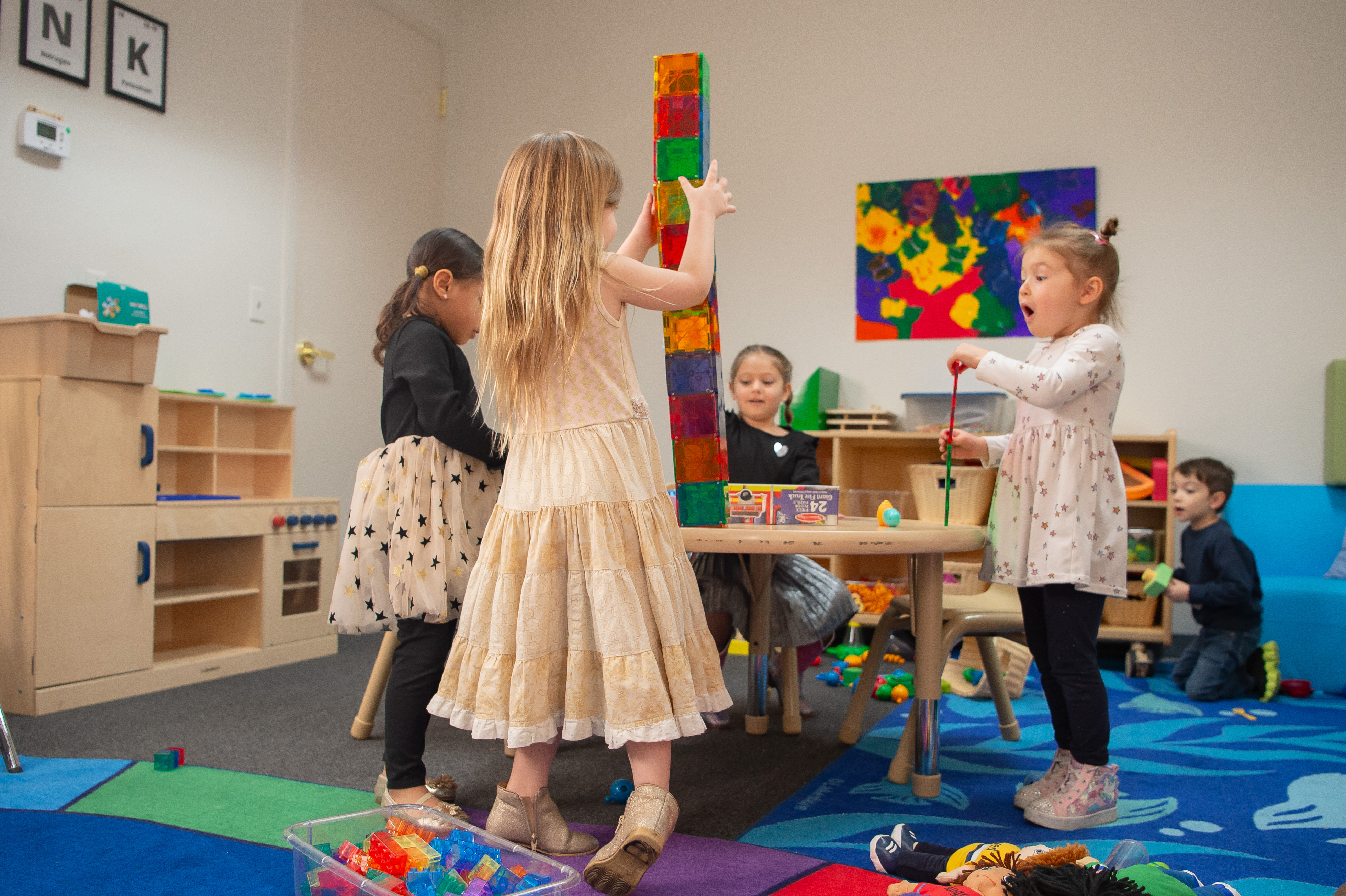 Group of 4 toddler girls play at a table with toys all over it. Laughing and smiling. 