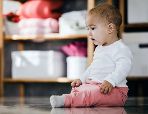 A baby with light skin and very little brown hair and her ears pierced is sitting up on the floor.