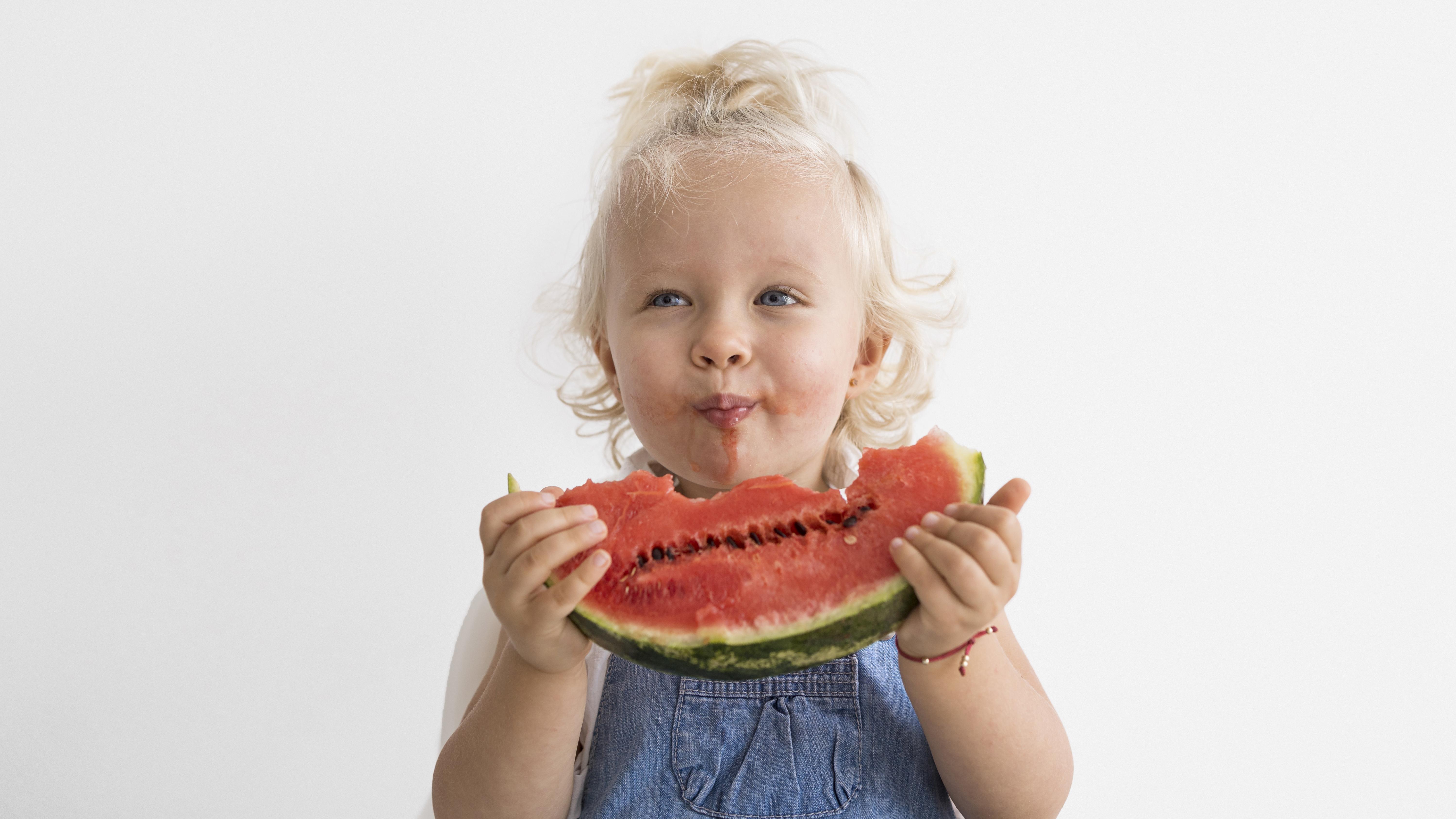 Toddler blonde girl wearing jean overalls, bites into a juicy watermelon with a big smile on her face.