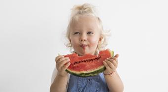Toddler blonde girl wearing jean overalls, bites into a juicy watermelon with a big smile on her face.