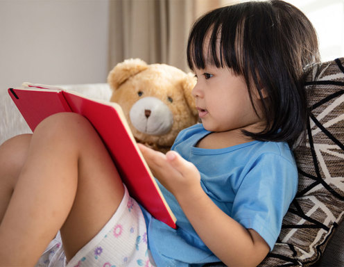 A young girl with light brown skin and straight dark hair reads a book to her teddy bear.