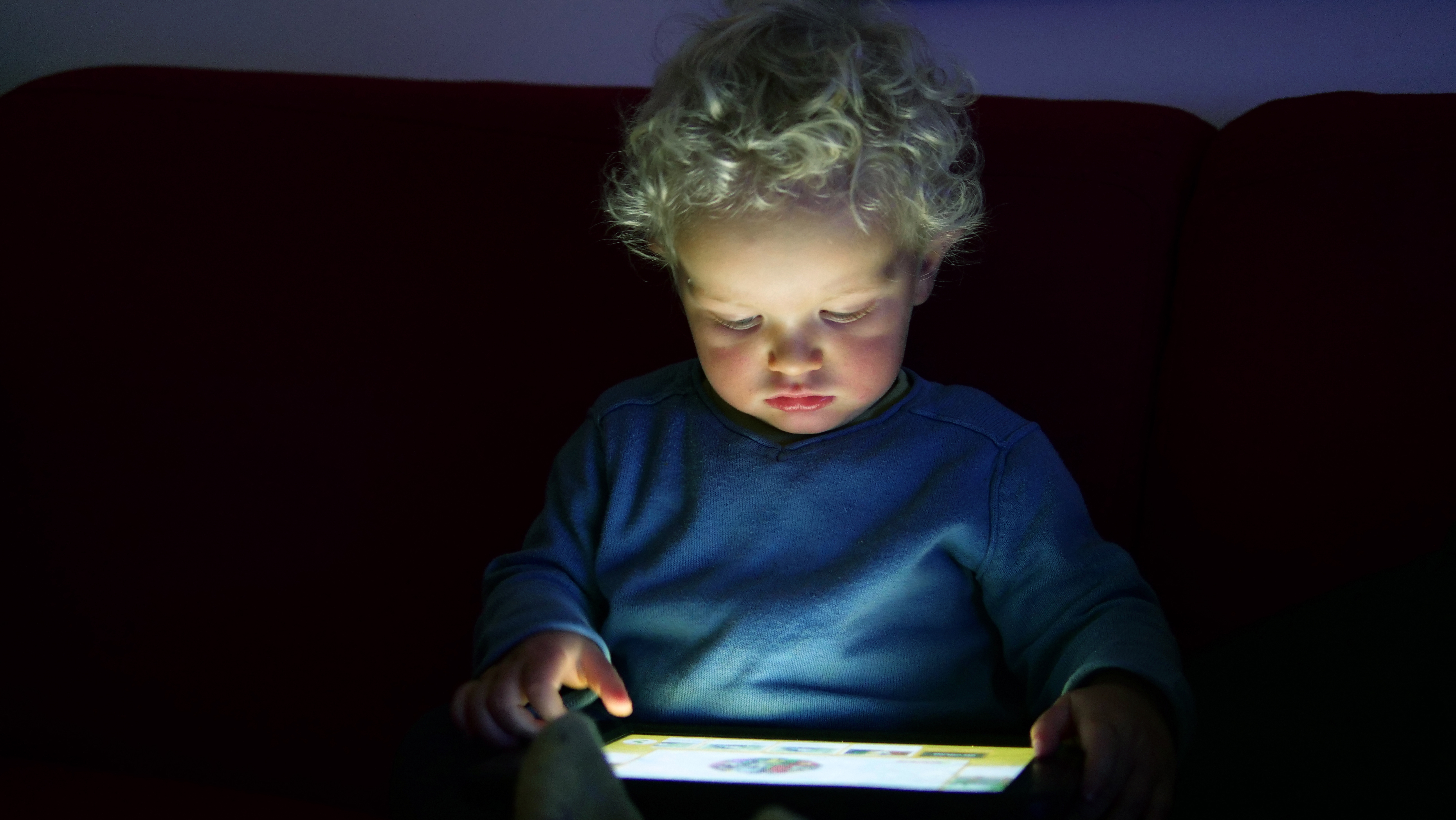 Young curly haired boy wearing blue shirt staring at I-pad screen in his lap in a low lighting setting 