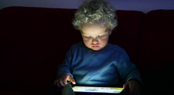 Young curly haired boy wearing blue shirt staring at I-pad screen in his lap in a low lighting setting 