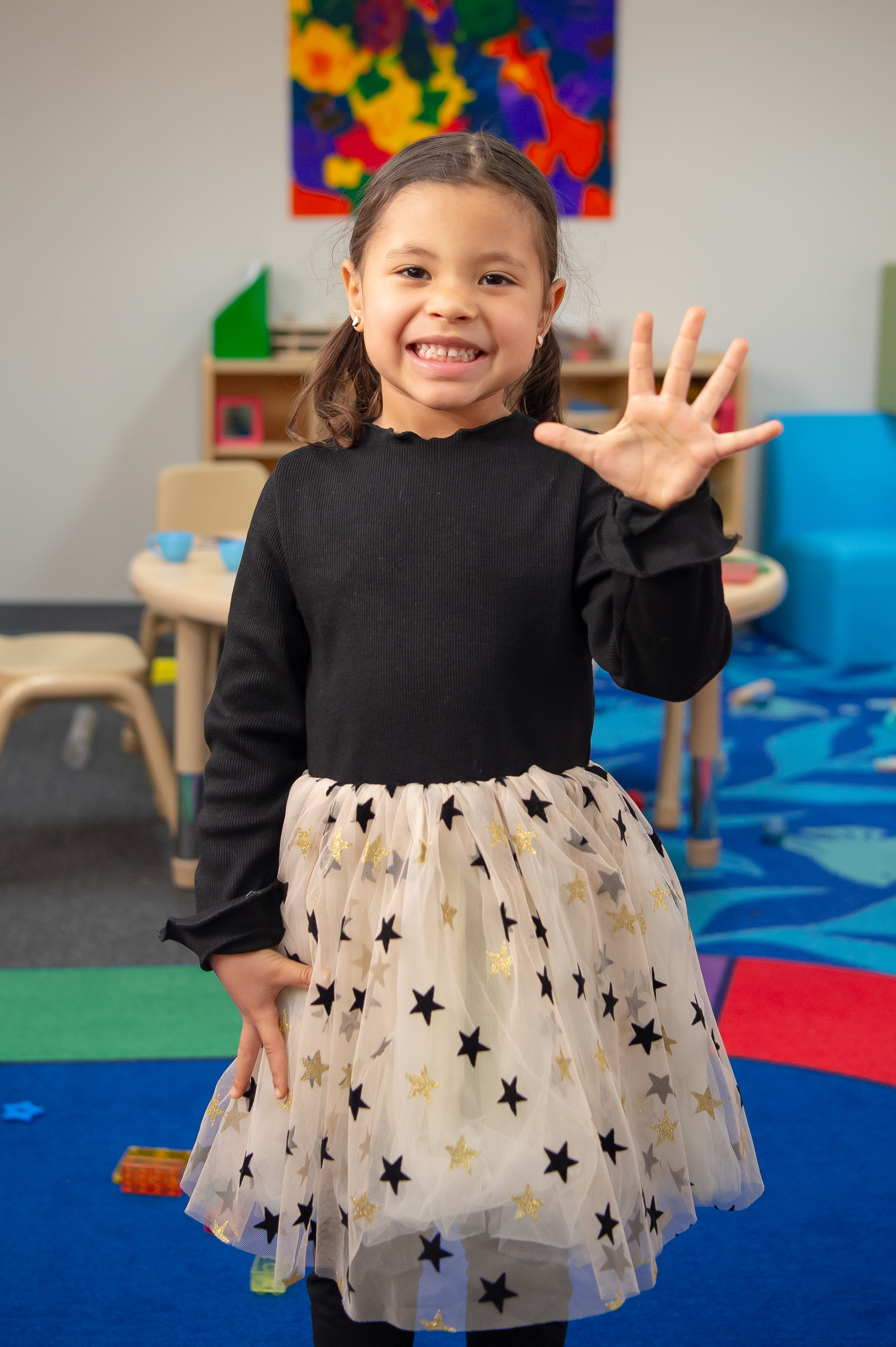 Smiling young girl in black long sleeved shirt with skirt covered in stars, looks at camera in a classroom setting