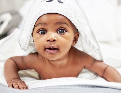 A baby with brown skin and brown eyes lying on its stomach holding its head up smiling at the camera with a bath towel lying over its head and body.