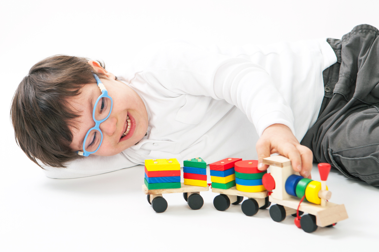 Young boy wearing blue glass with light skin and short brown hair laying on the floor playing with a train toy. 