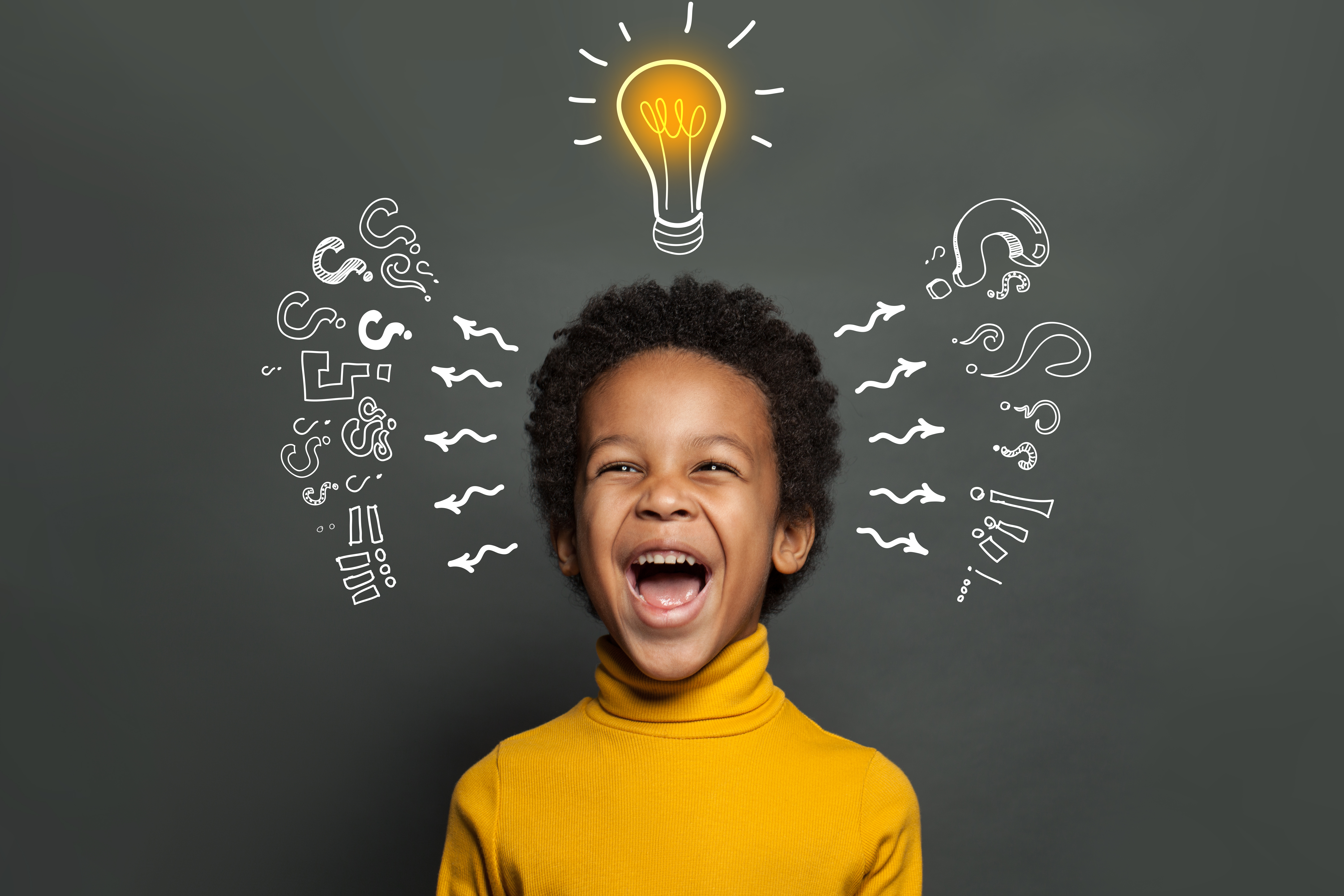 An African-American young boy with short curly hair wearing a bright yellow shirt, smiling and standing in front of a chalkboard with a light bulb drawn in chalk above his head.