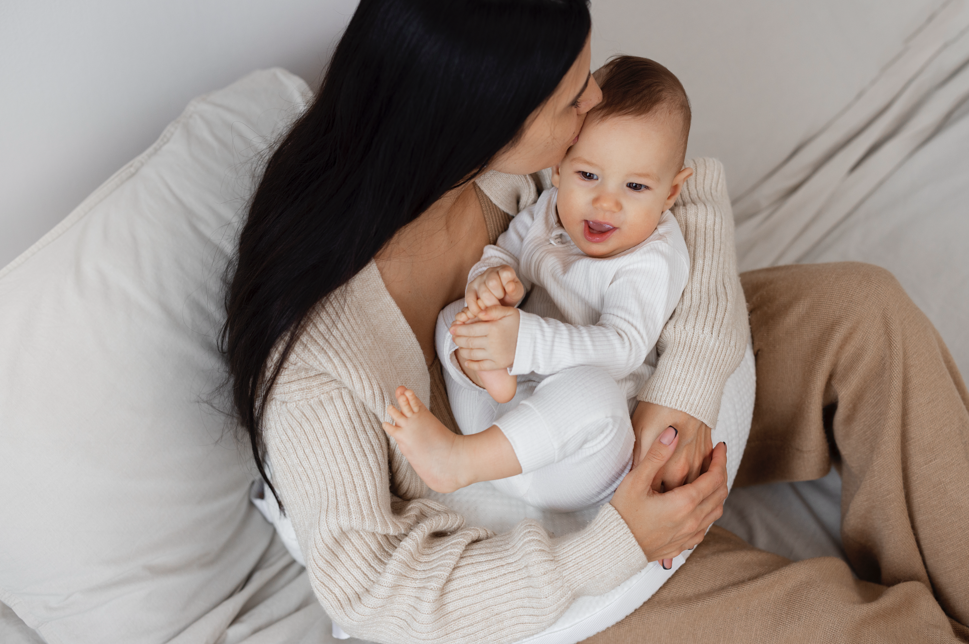 Dark haired women sitting on comfy couch with a baby smiling in her lap. 