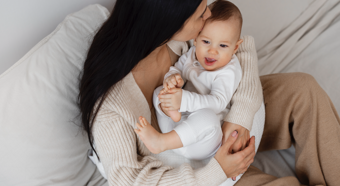 Dark haired women sitting on comfy couch with a baby smiling in her lap. 