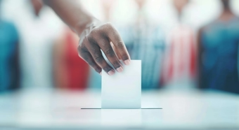 A hand in front of a blurry voting area background. Hand is casting a vote into a voting box