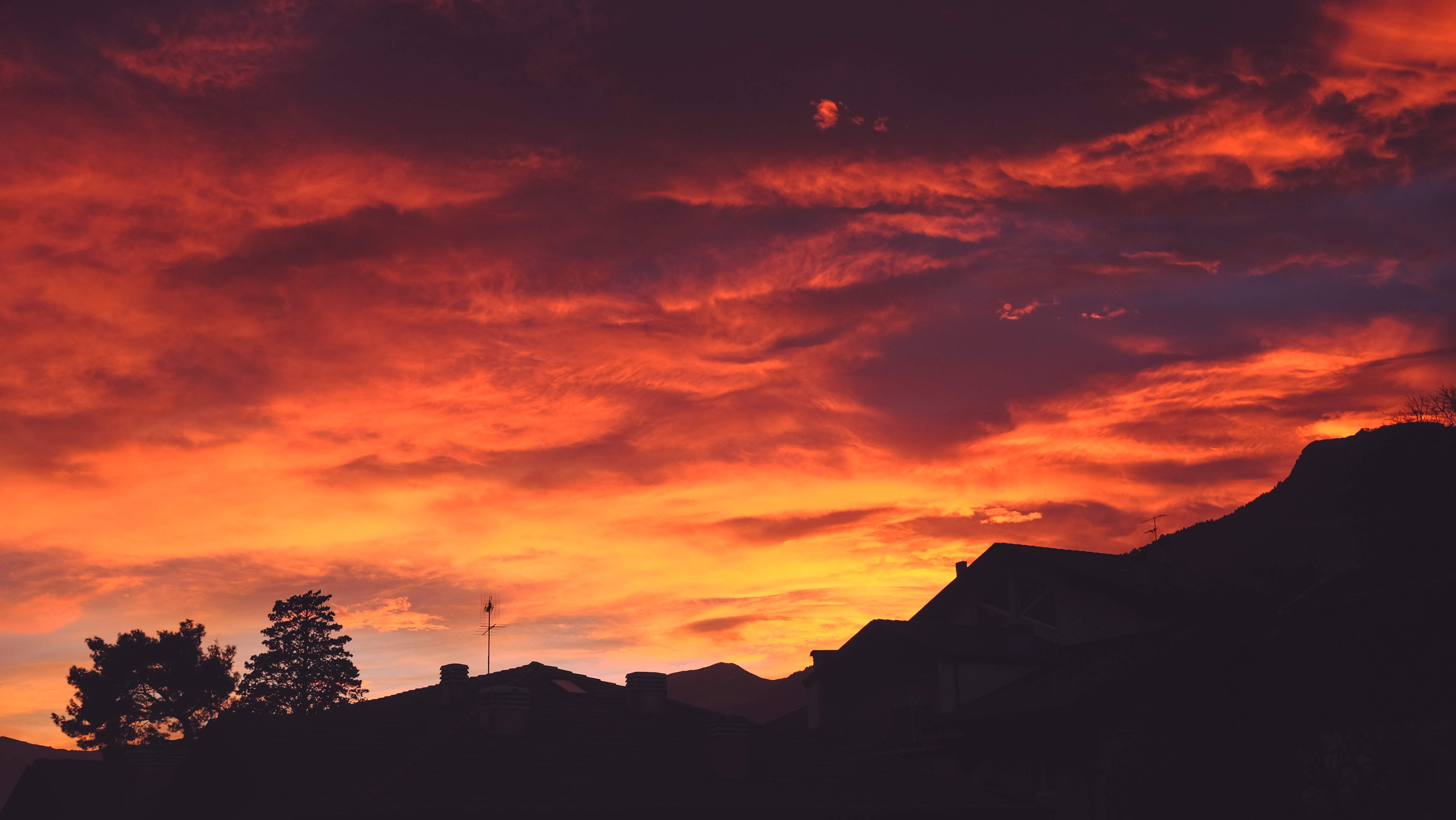 Sunset sky with shades of red, orange and yellow over a neighborhood with mountains and trees in the foreground and background 