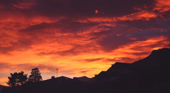 Sunset sky with shades of red, orange and yellow over a neighborhood with mountains and trees in the foreground and background 