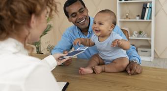 Baby boy smiling and laughing, sitting on a desk with adults smiling at table 