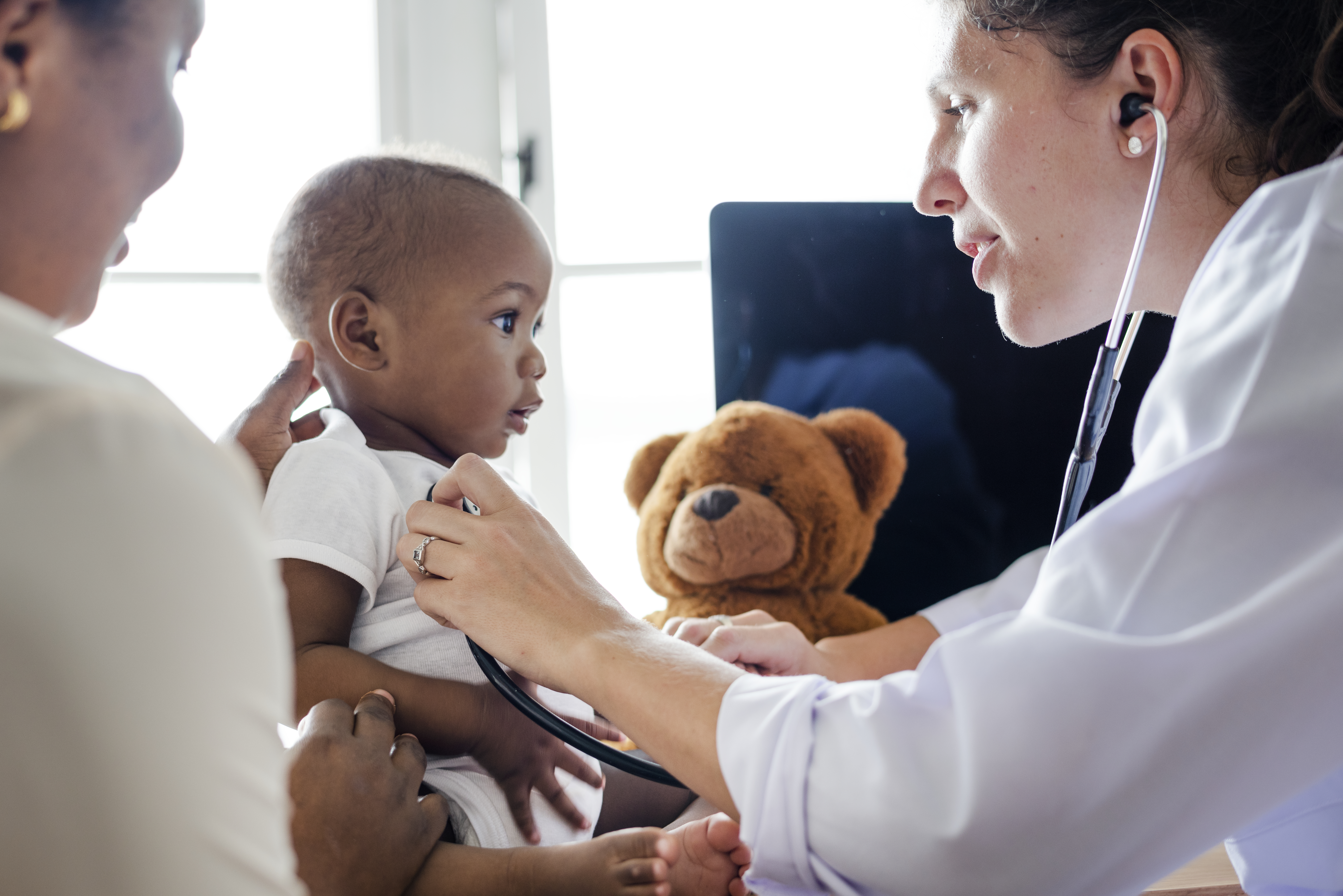 Women in a doctors office setting checking a little baby boys heart, with another adult figure sitting with the child