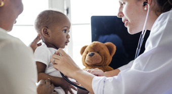 Women in a doctors office setting checking a little baby boys heart, with another adult figure sitting with the child