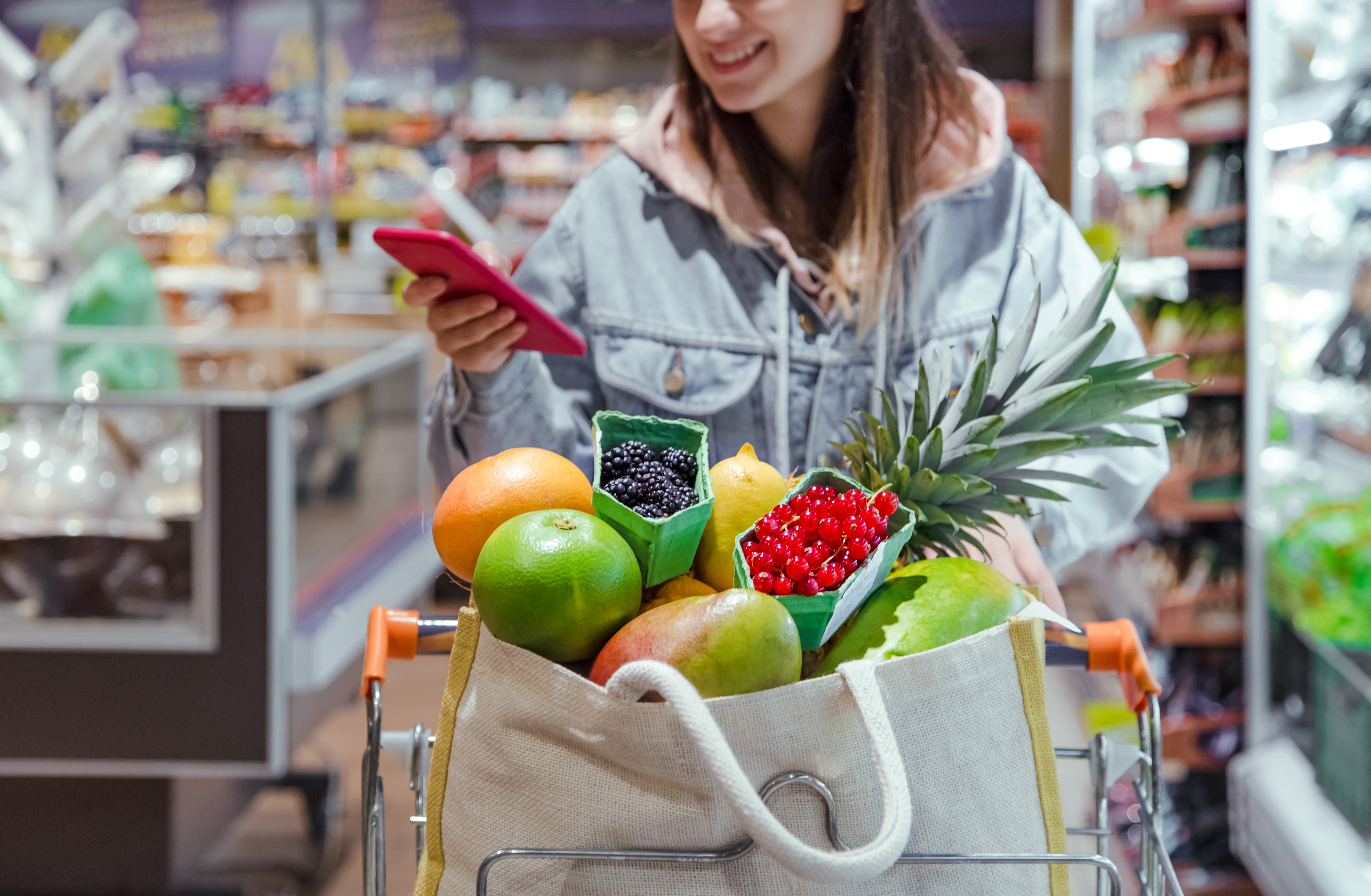 Young women with grocery cart full of fruits and vegetables, looking down at her phone while smiling 