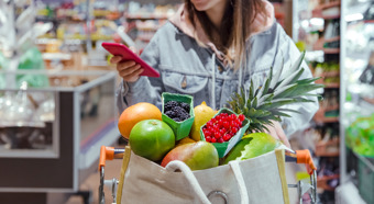 Young women with grocery cart full of fruits and vegetables, looking down at her phone while smiling 