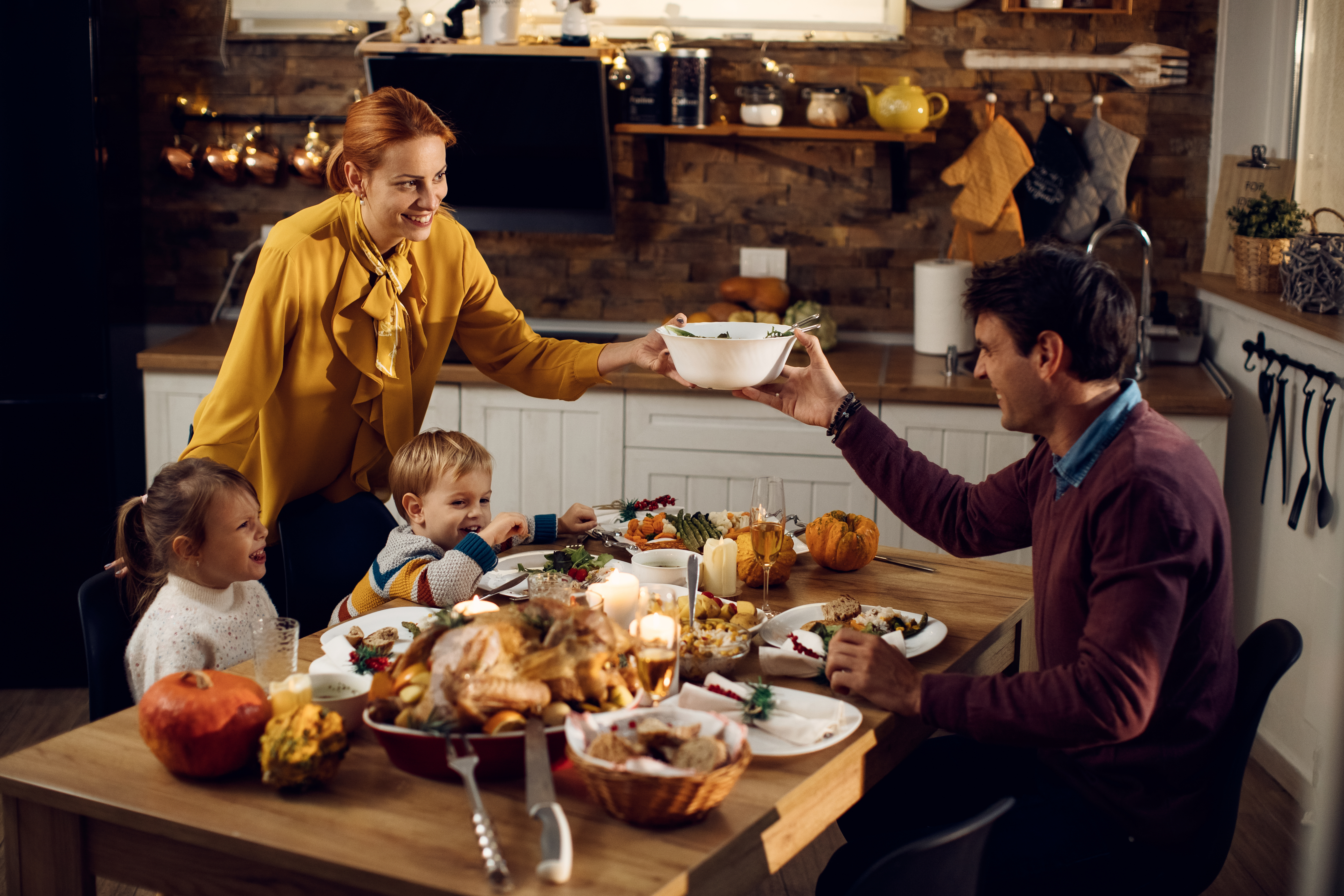 Thanksgiving dinner table with thanksgiving foods covering the table, young boy, girl, and two adults sitting around the table laughing and smiling