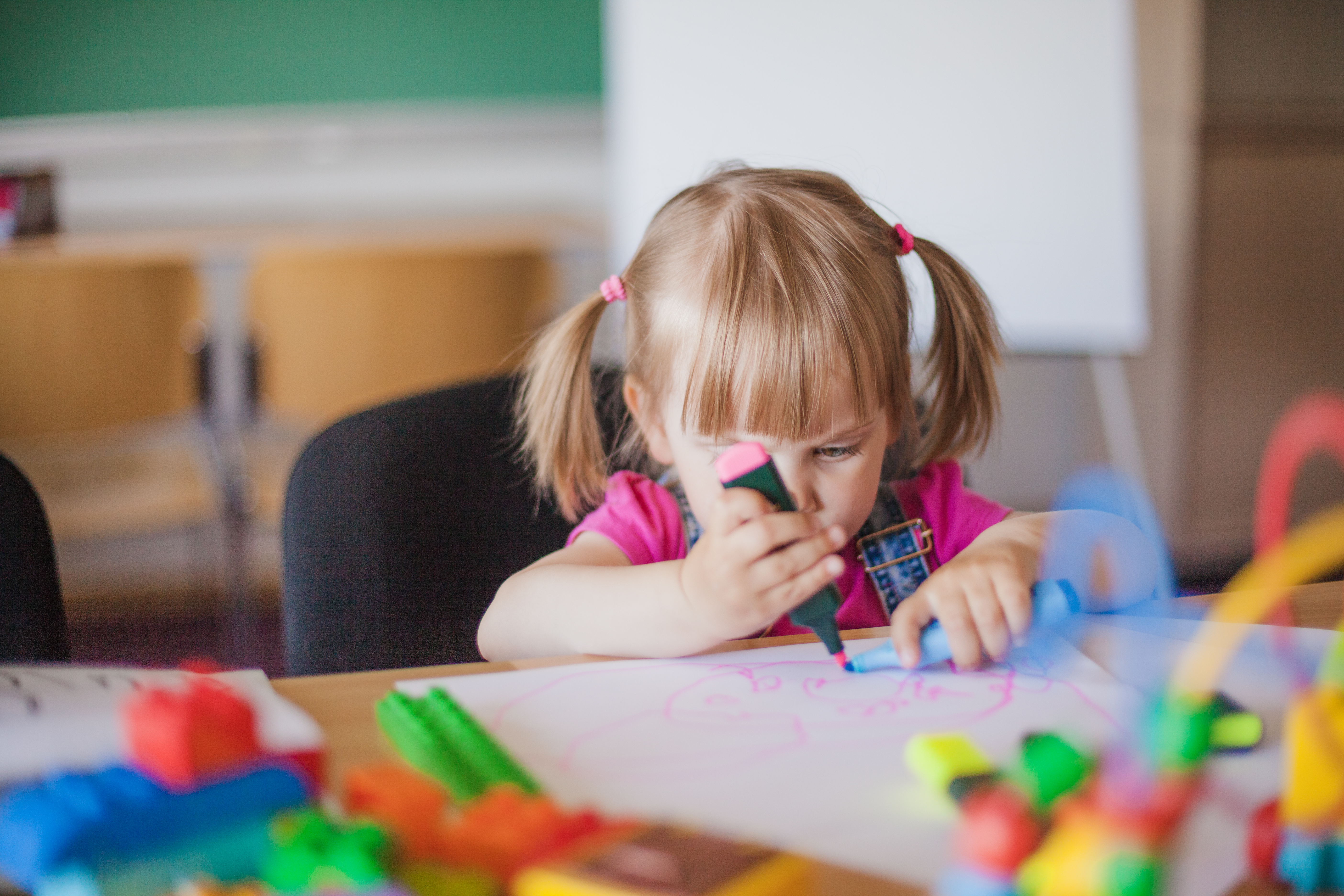 Little girl draws on piece of paper with pink marker in a school setting 