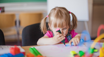 Little girl draws on piece of paper with pink marker in a school setting 