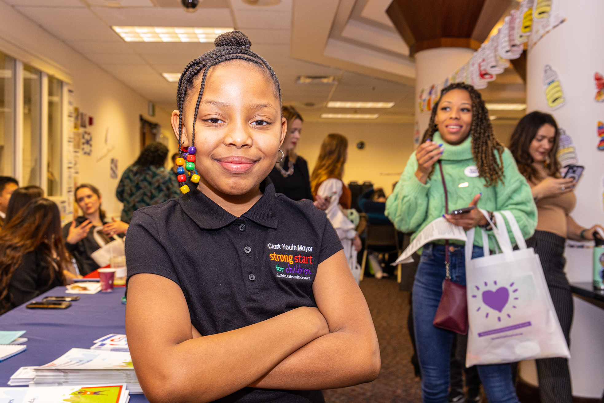 A young African American girl with braided hair and beads stands looking at the camera smiling with her arms crossed with adults and tables behind her.