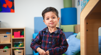 Young boy with plaid shirt smiling and looking at camera for photo in a classroom setting.