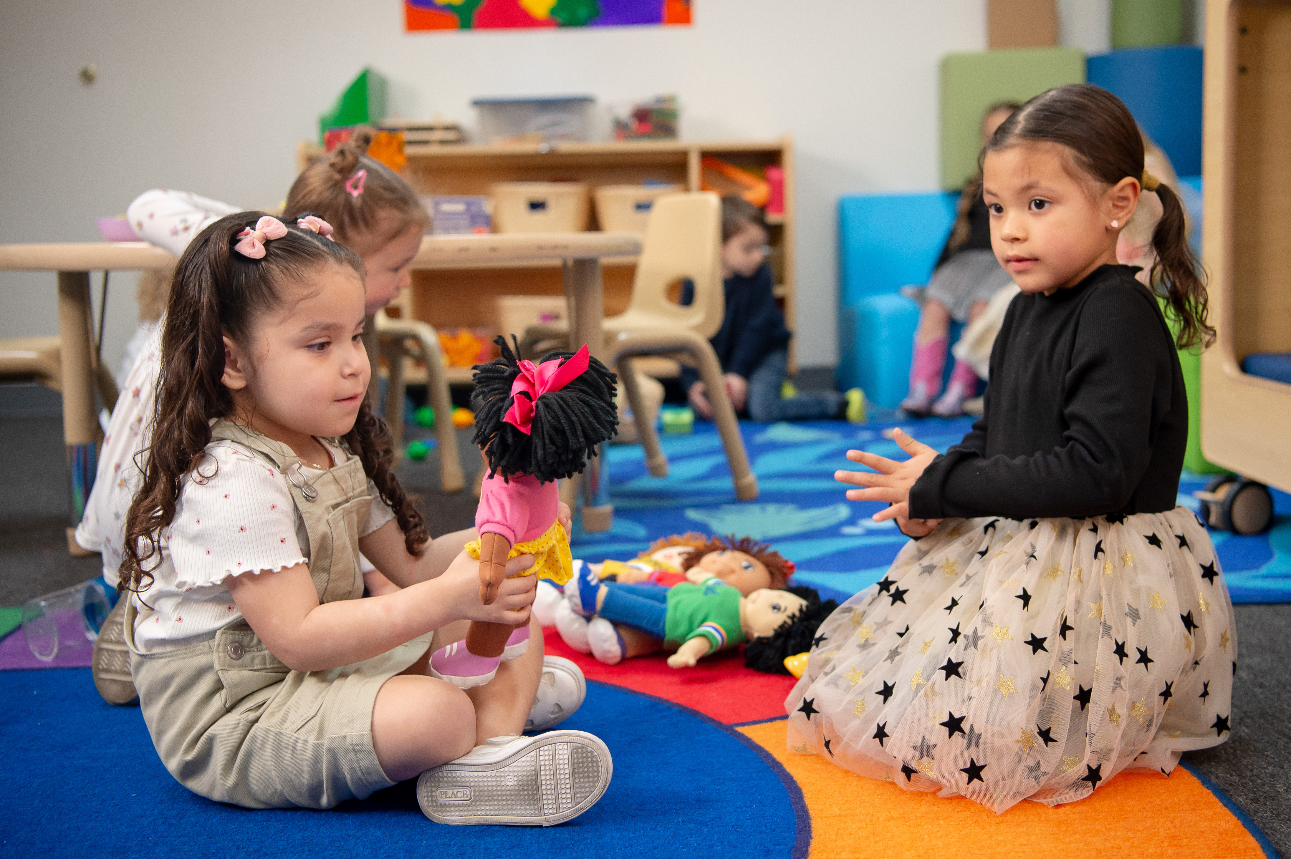 Two hispanic girls sitting on a colorful carpeted floor and Playing with dolls. 