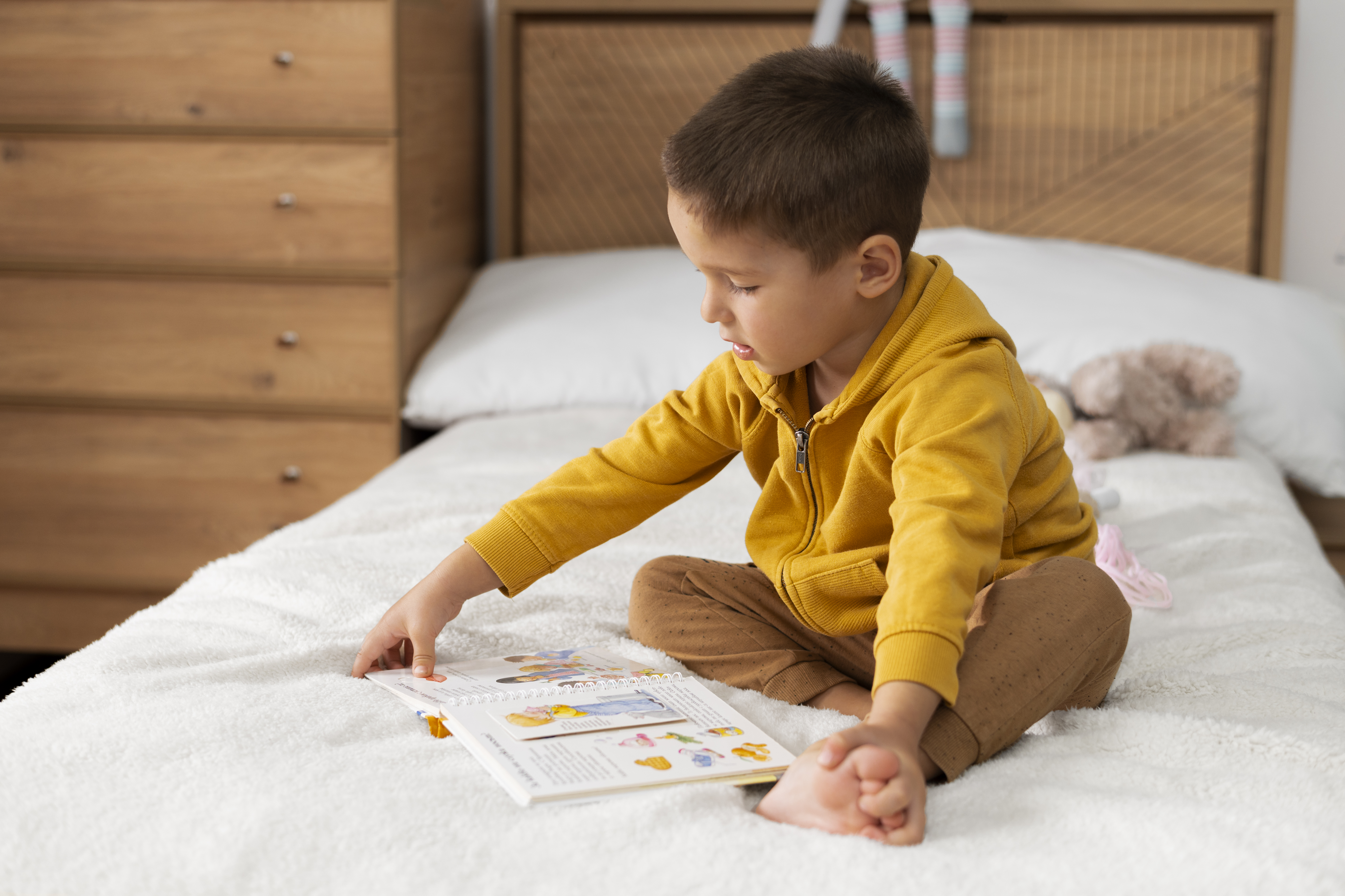 Toddler boy wearing yellow long-sleeved shirt, sits on bed setting while reading a children's book. 