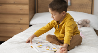 Toddler boy wearing yellow long-sleeved shirt, sits on bed setting while reading a children's book. 