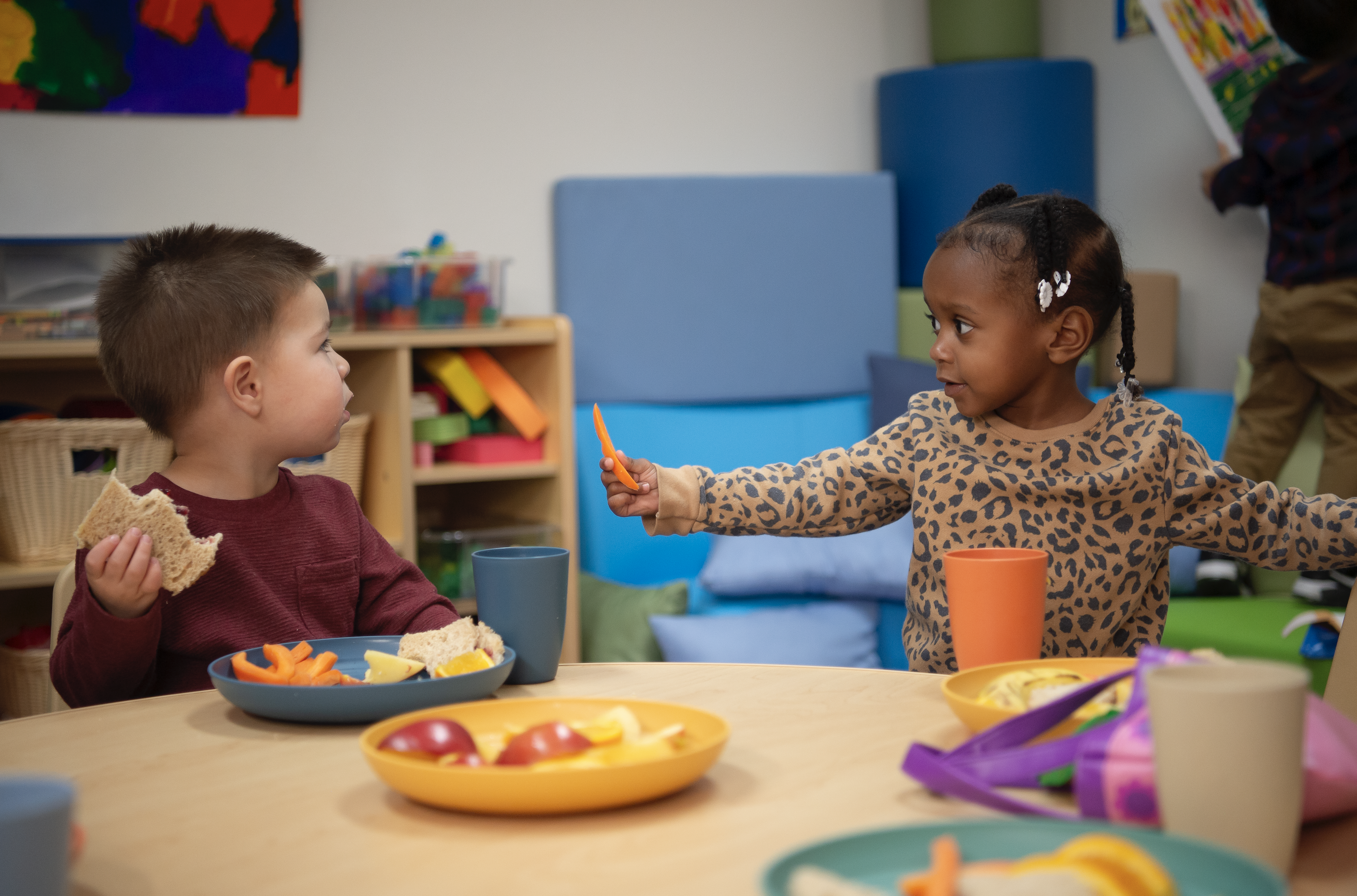 Young girl holding vegetable at table with young boy eating a sandwich, in a classroom setting