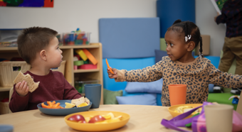 Young girl holding vegetable at table with young boy eating a sandwich, in a classroom setting