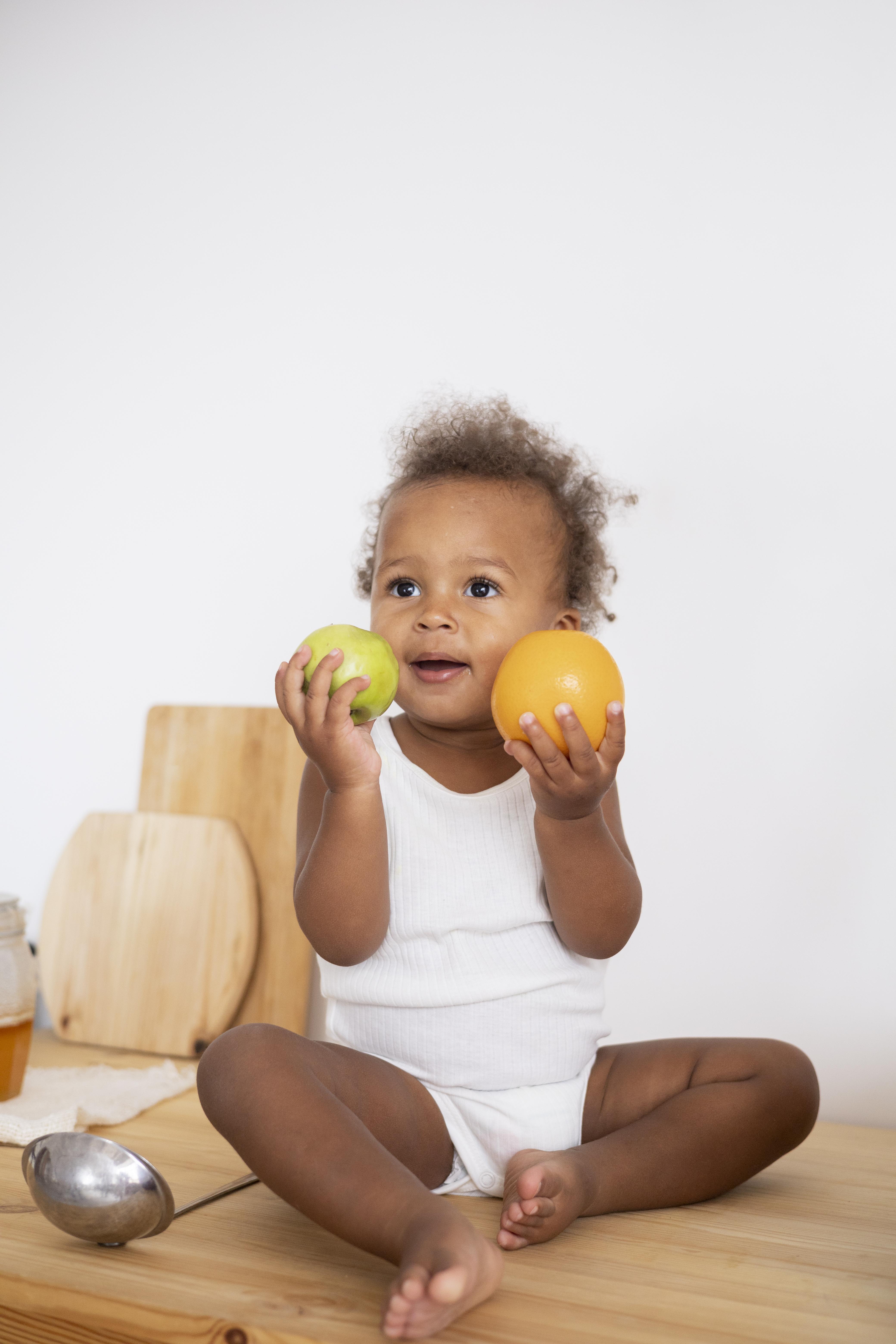 Young toddler sitting on kitchen counter setting, wearing all white, holds two fruits in hands while smiling and looking off into the distance. 