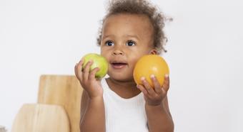 Young toddler sitting on kitchen counter setting, wearing all white, holds two fruits in hands while smiling and looking off into the distance. 