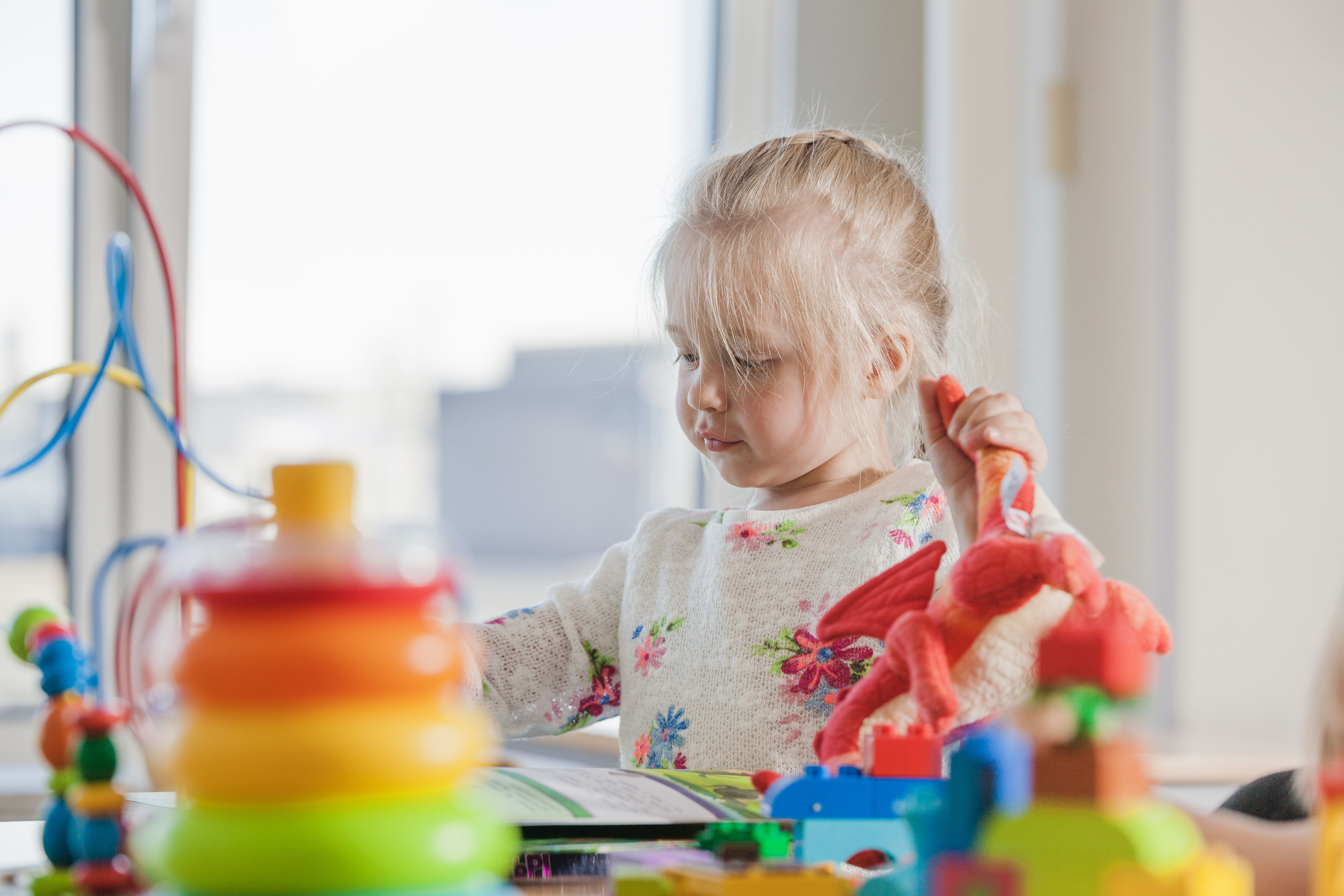 Young blonde toddler wearing white shirt with flowers, playing at table full of colorful toys. 