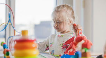 Young blonde toddler wearing white shirt with flowers, playing at table full of colorful toys. 