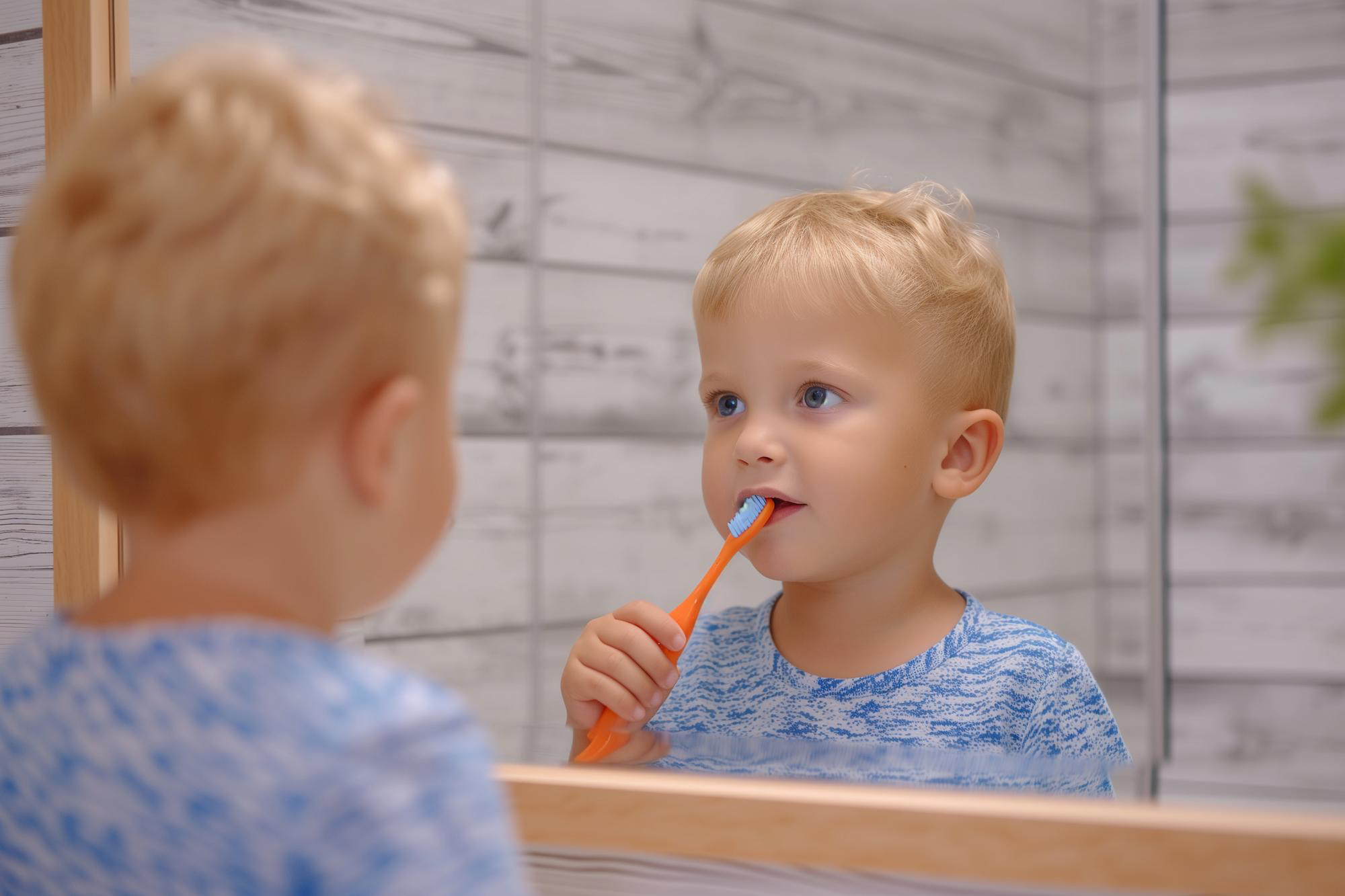 Baby boy with blonde hair stares at reflection in mirror while brushing his teeth with orange toothbrush 