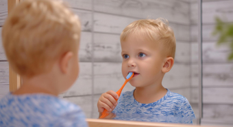 Baby boy with blonde hair stares at reflection in mirror while brushing his teeth with orange toothbrush 