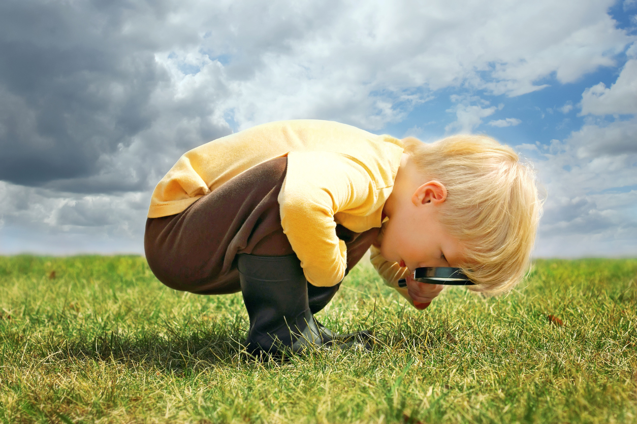 A toddler-aged boy with light skin and blonde hair is bending over looking at the grass with a magnifying glass.